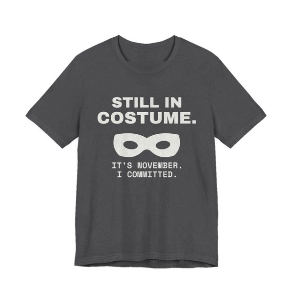 Dark Grey Heather t-shirt printed in off-white ink with “Still in Costume.” above an eye mask, with “It's November. I committed.”