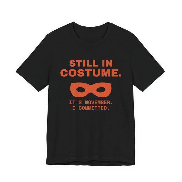 Black t-shirt printed in orange ink with “Still in Costume.” above an eye mask, with “It's November. I committed.”