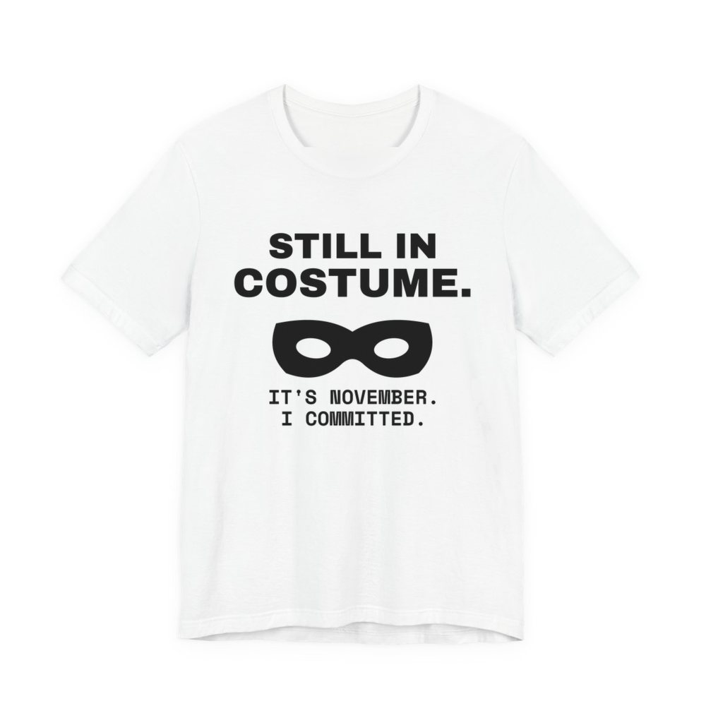 White t-shirt printed in dark ink with “Still in Costume.” above an eye mask, with “It's November. I committed.”